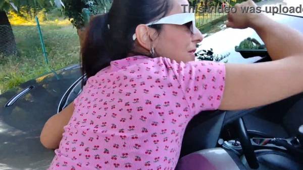 Young woman in pink dress gives bold outdoor blowjob beside a car while someone watches on adultfans.net