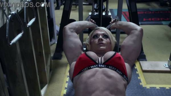 Muscular blonde flexes her defined physique and full breasts in a dim gym on adultfans.net